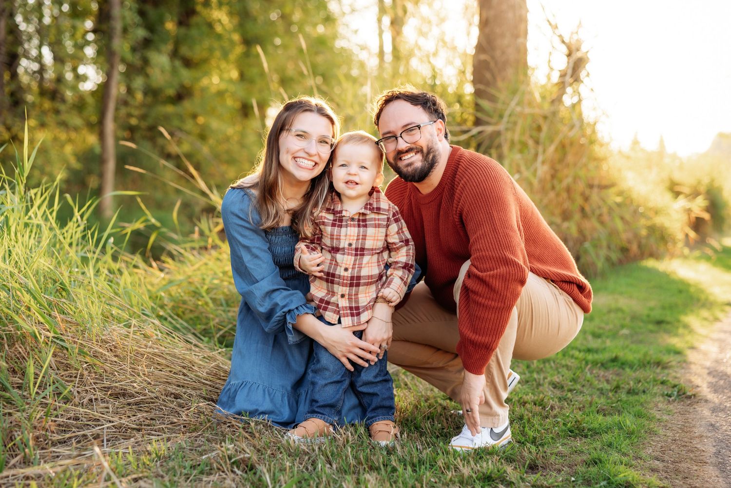 Happy family of three posing outdoors in golden hour light, parents smiling with toddler on a scenic nature path.