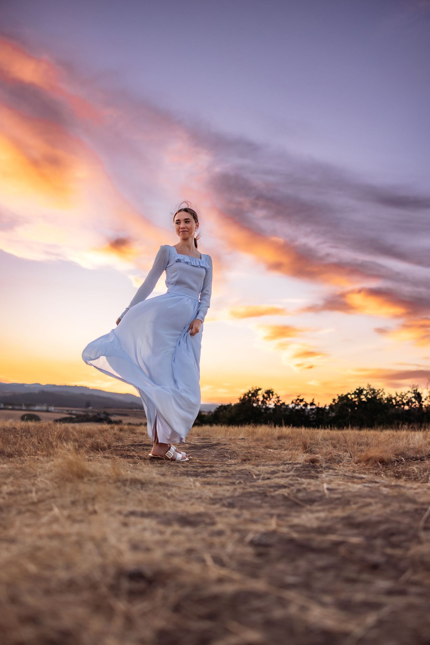 Woman in flowing blue dress stands in golden field at sunset with dramatic purple and orange sky.