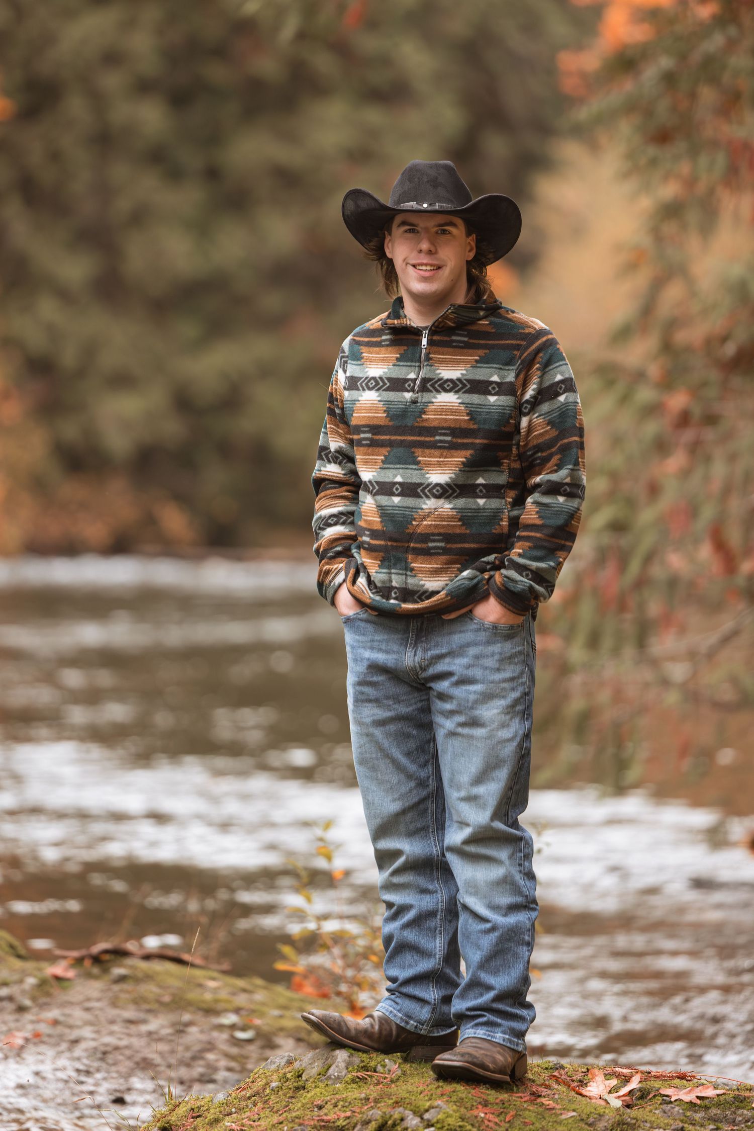 A small child wearing a patterned sweater and cowboy hat stands near a stream in an autumn woodland setting.