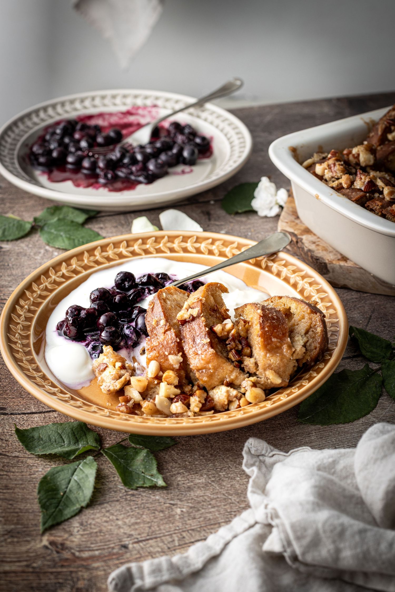 Baked French toast served on a plate with smashed blueberries and Greek yoghurt, and a plate of blueberries on the table in the background.