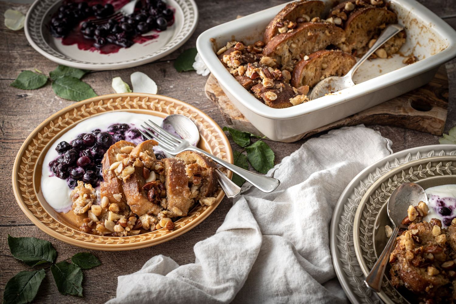 A table laid with plates and a serving dish containing baked French toast, also served on a plate with smashed blueberries and Greek yoghurt