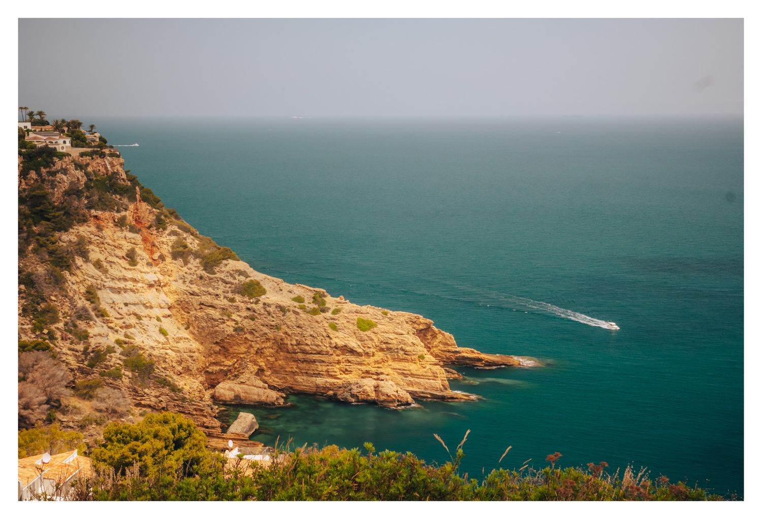 Rugged coastal cliffs meet turquoise Mediterranean waters with a small boat visible in the distance.