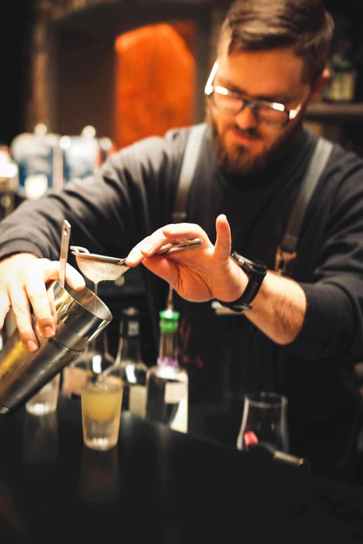 A bartender in glasses pours a drink from a metal shaker at a dimly lit bar counter.