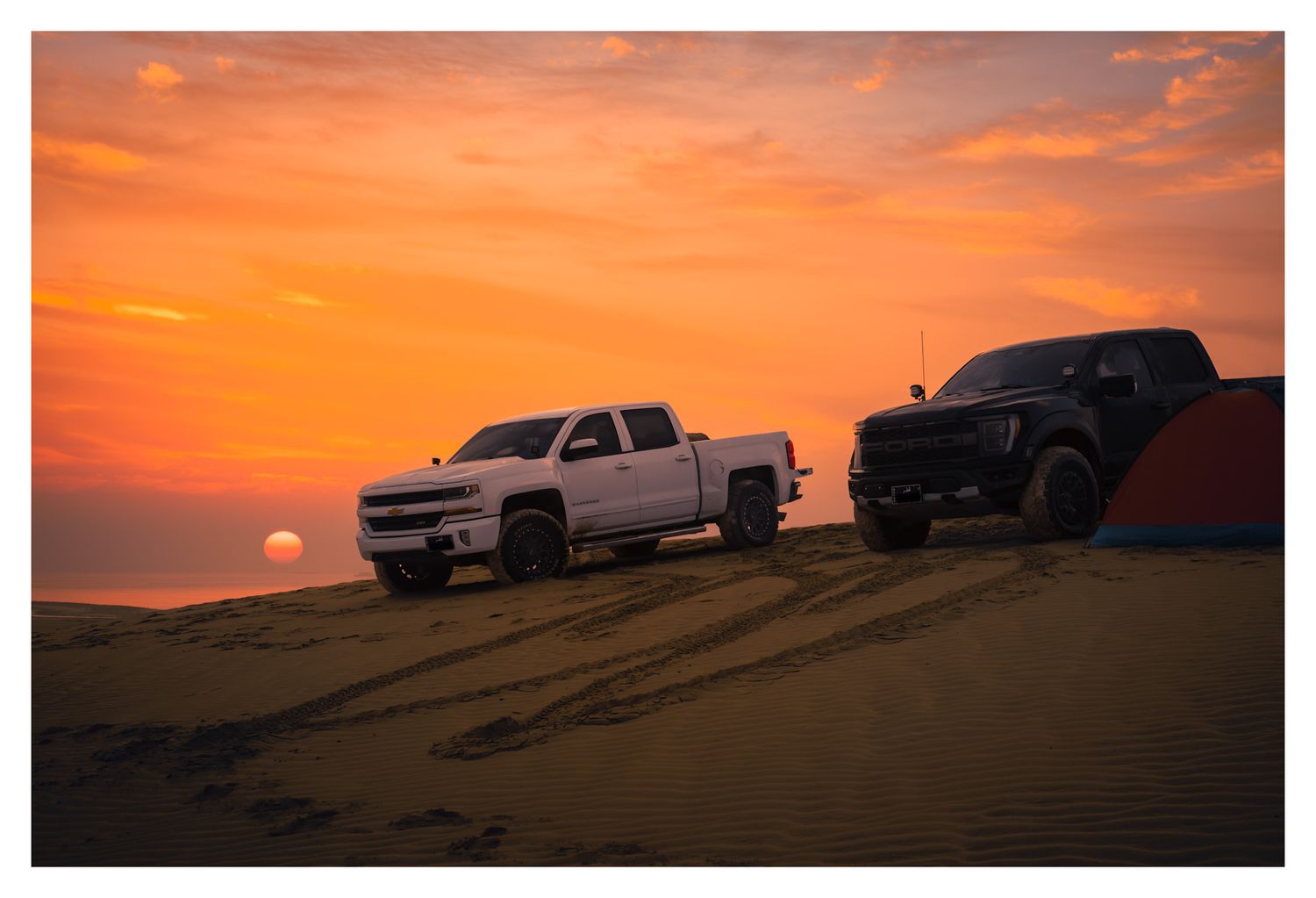 Pickup trucks silhouetted against a vibrant orange sunset sky over desert sand dunes.