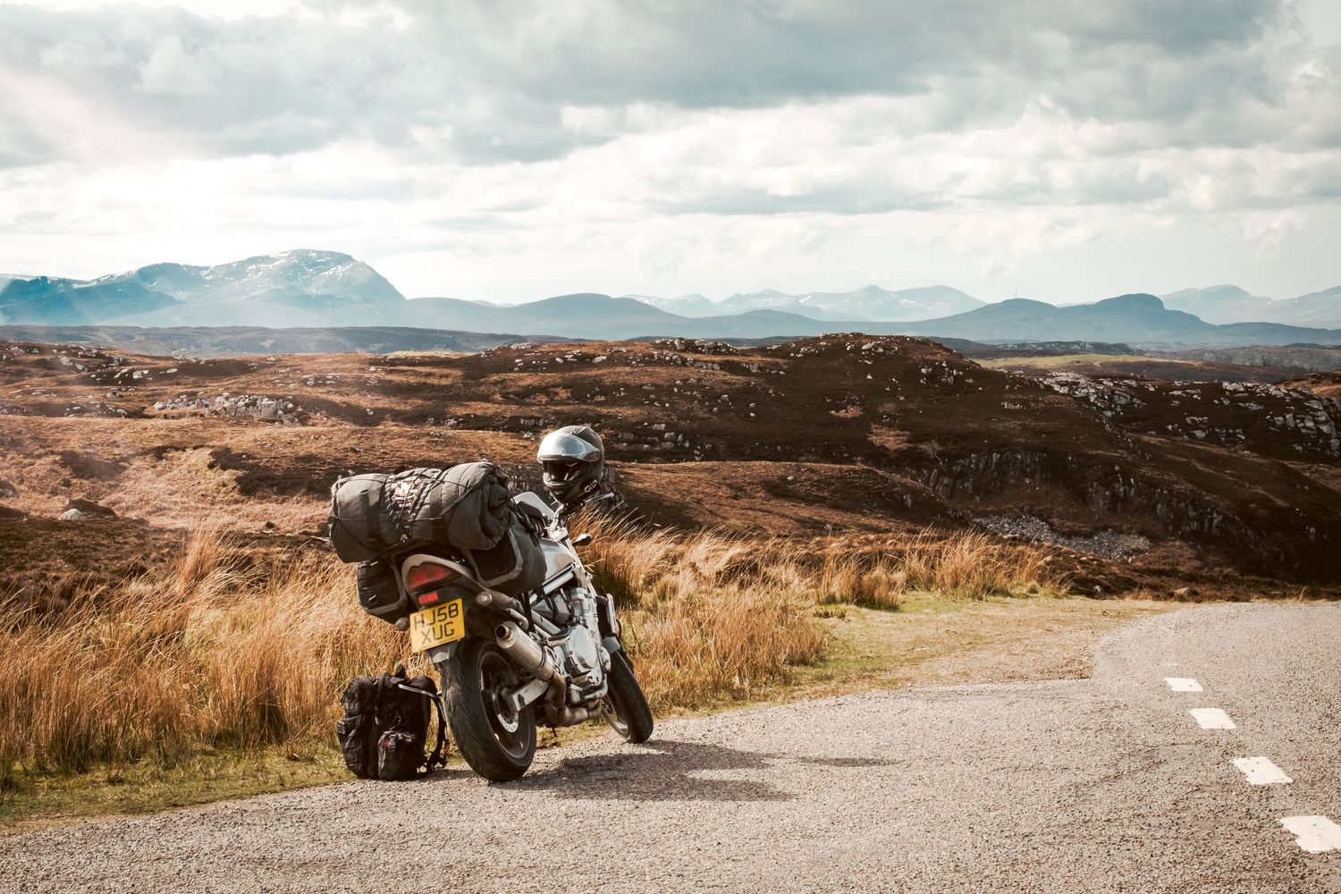 Loaded adventure motorcycle parked on scenic highland road with rugged mountains and dramatic cloudy sky in background.