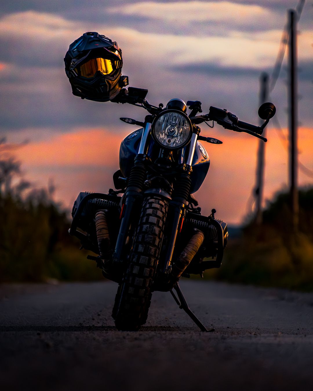 A motorcycle with black helmet rests on a road against a dramatic sunset sky with orange and purple hues.
