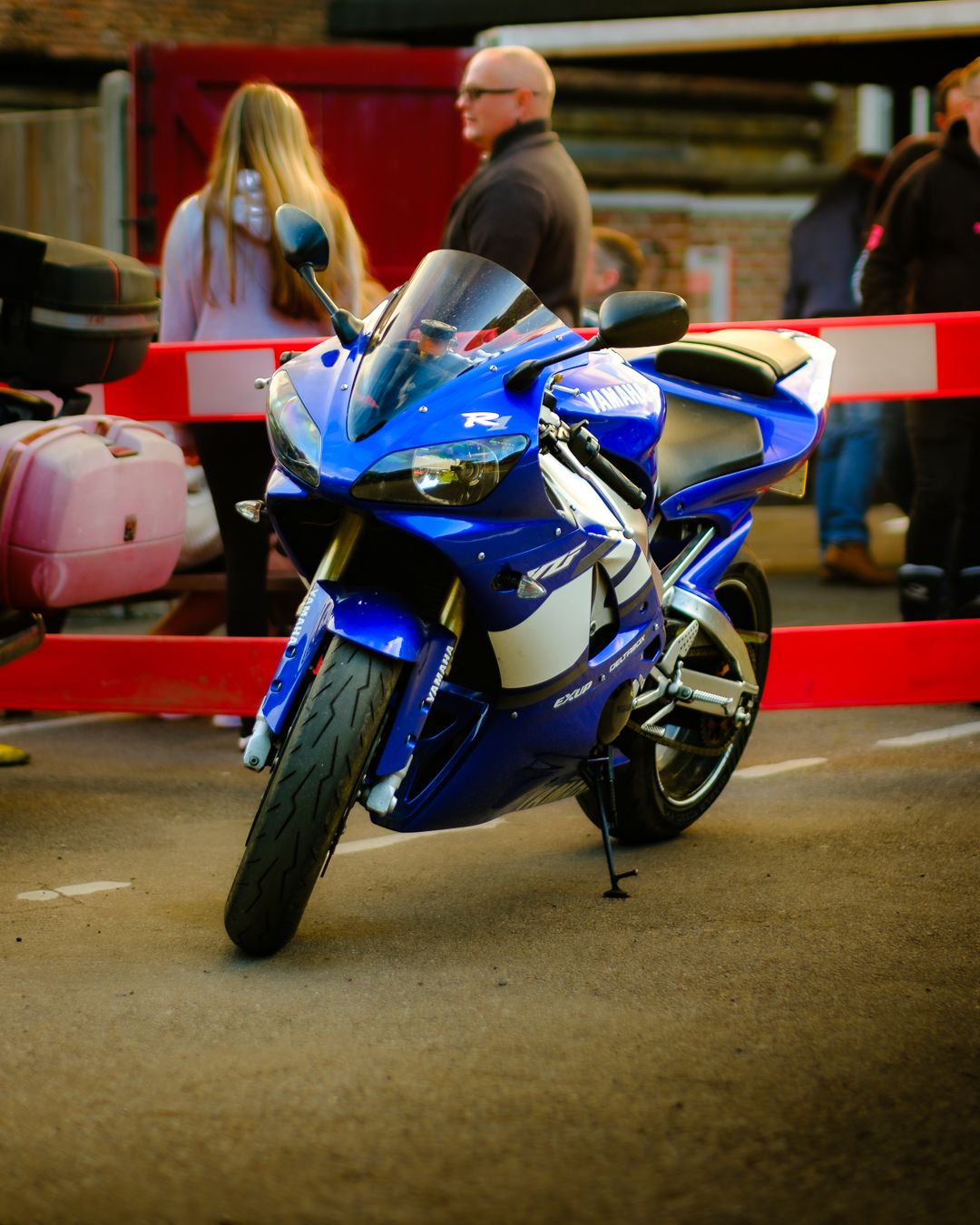 A blue and white sport motorcycle parked at an outdoor location with red barriers visible in the background.