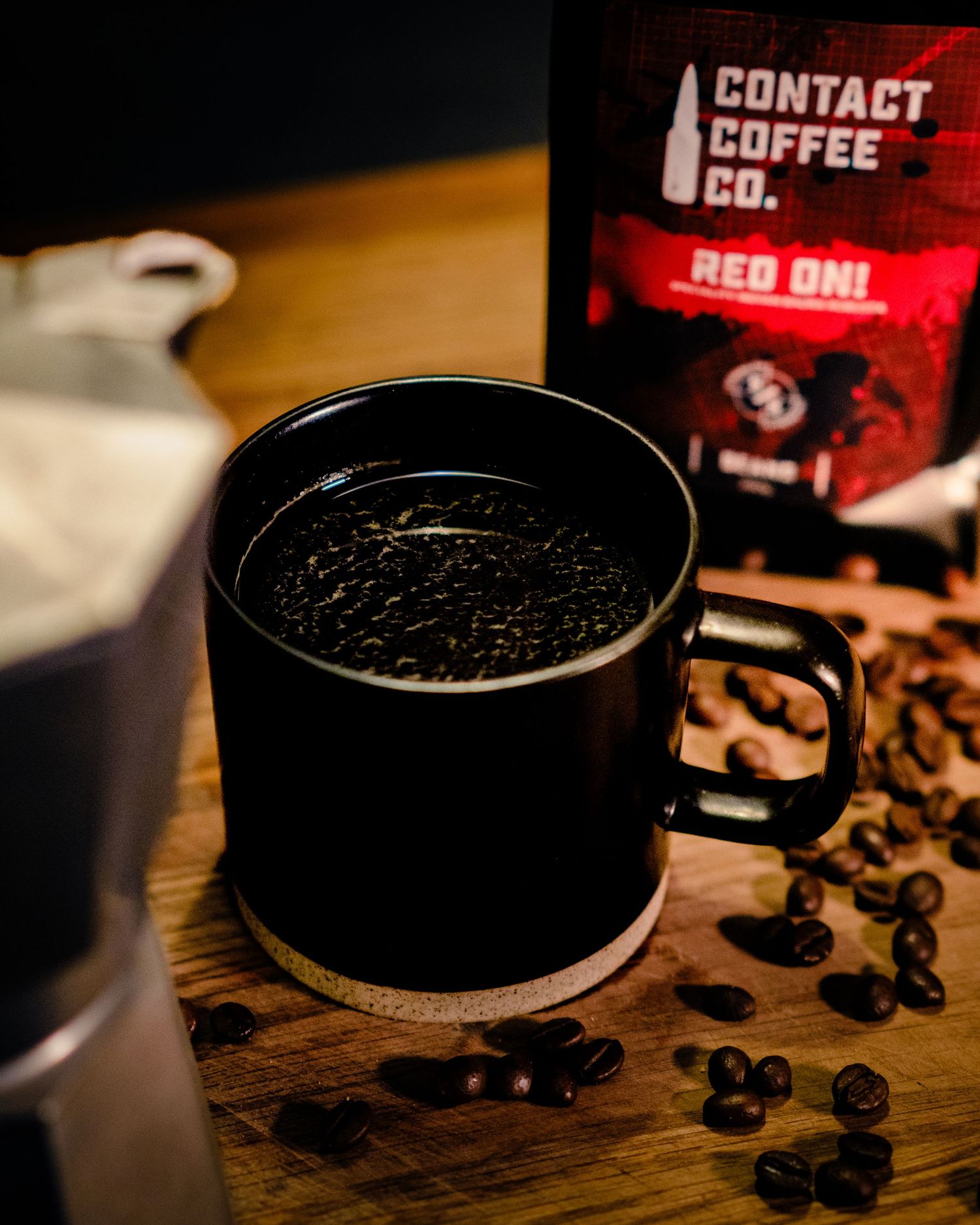 A black coffee mug with dark roasted beans scattered on a wooden table next to a Contact Coffee Co Red On coffee bag.