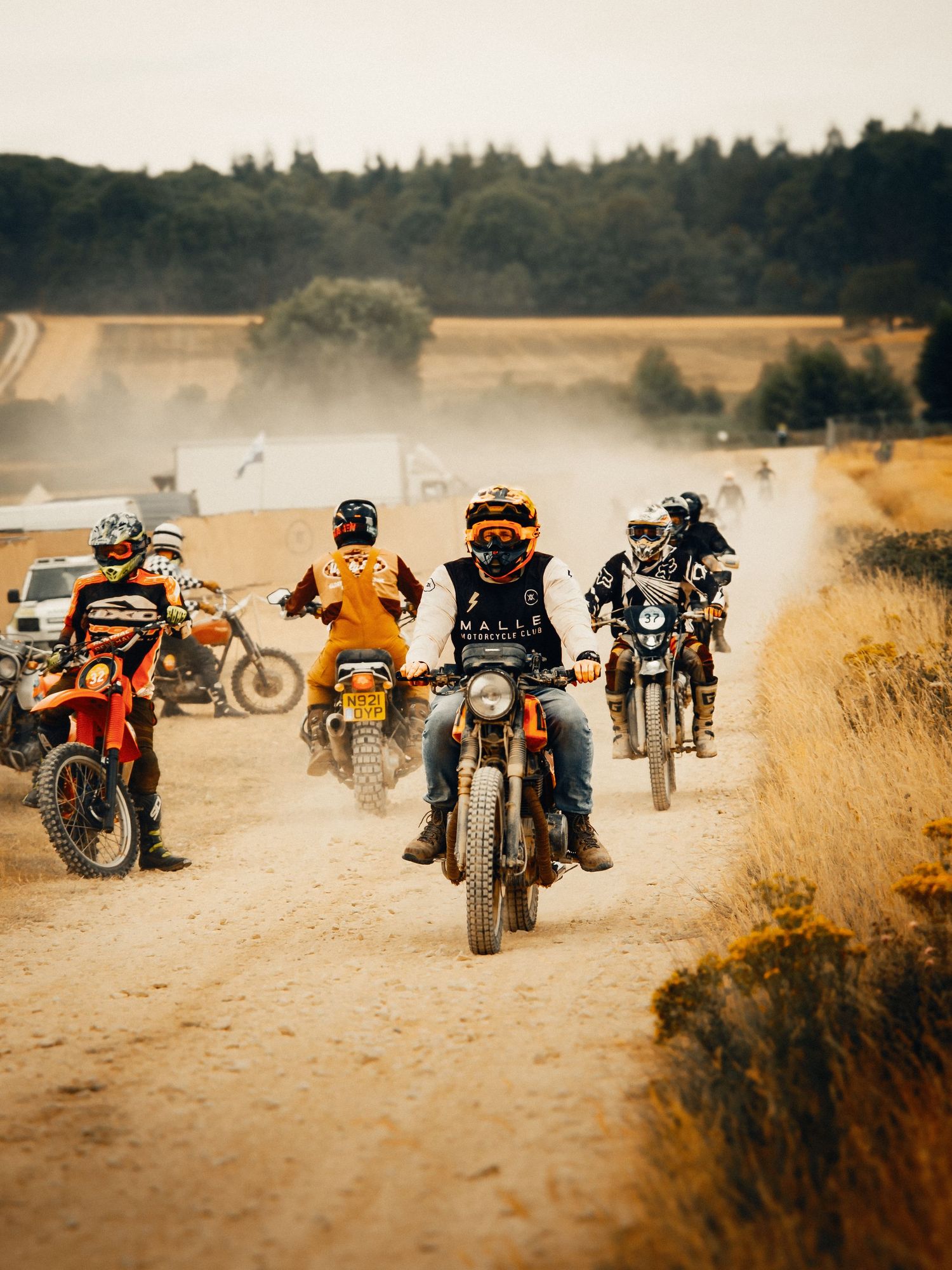Group of motocross riders racing on a dusty dirt trail with trees and fields in the background.
