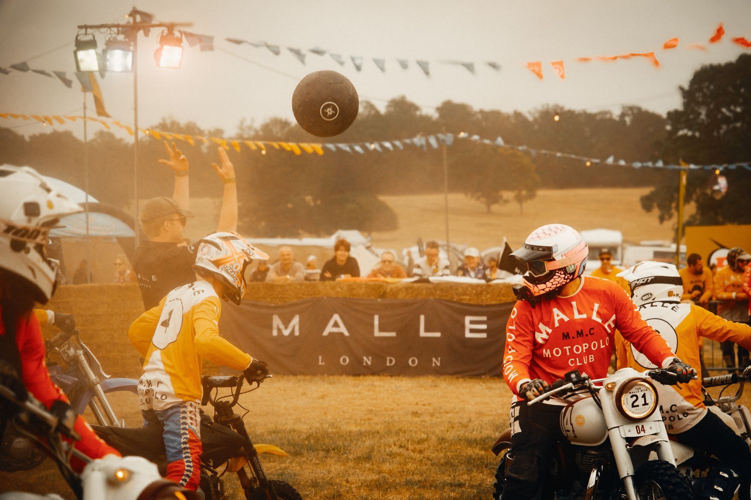 Motorcyclists in retro gear at Malle London outdoor moto festival with banner and crowd in background.