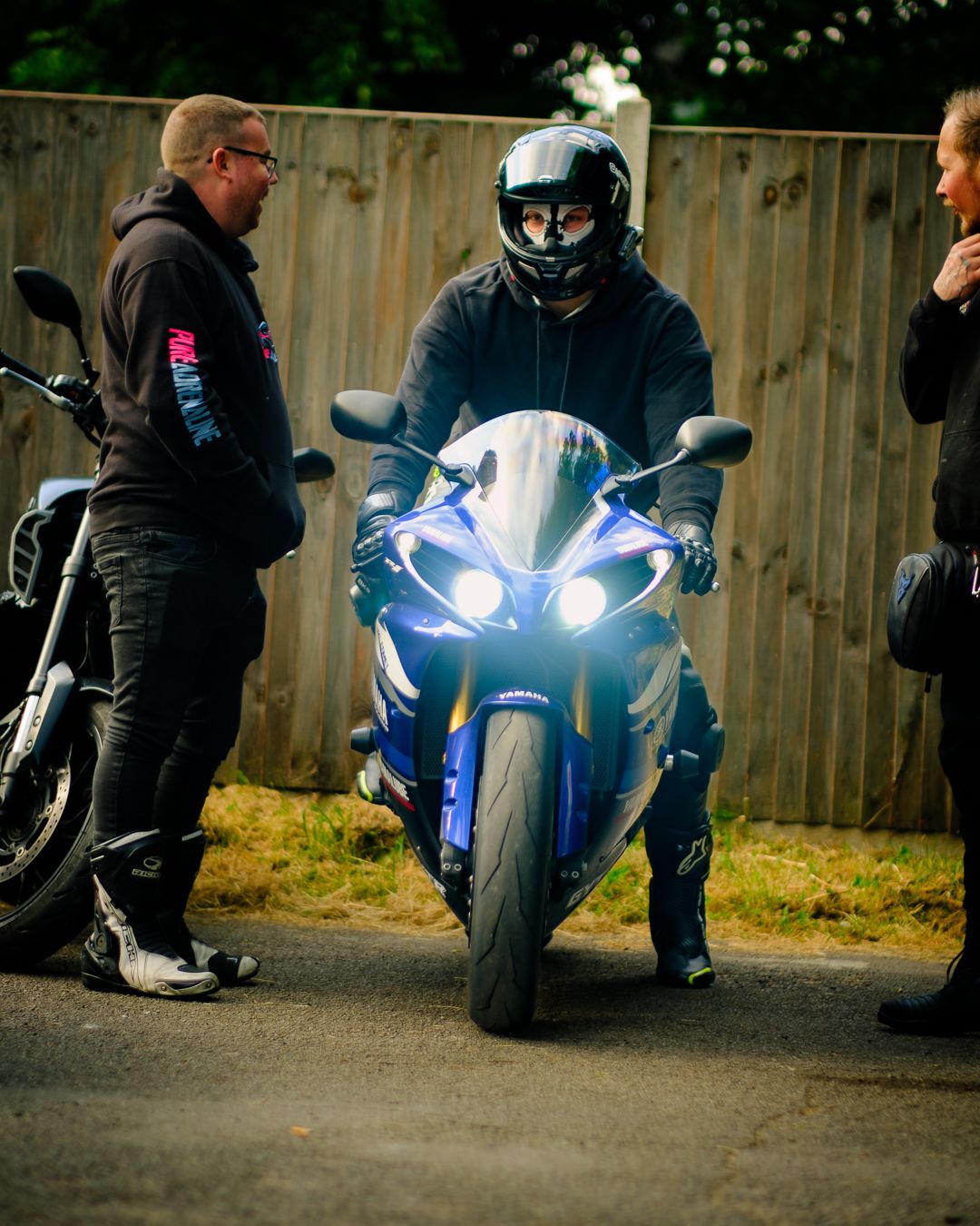 A series of motorcycles parked at night with bright LED headlights illuminating the darkness against a wooden fence background.