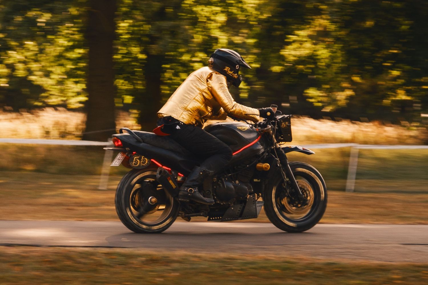 Person in yellow jacket and helmet riding a black motorcycle on a road with blurred green trees in the background.