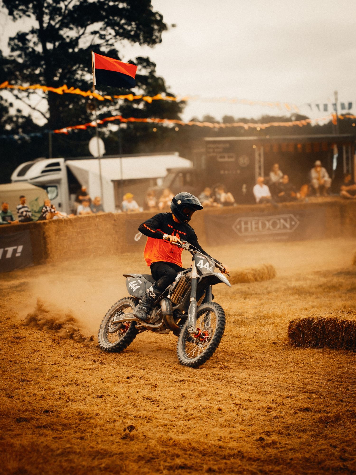 A motocross rider in orange gear races through a dusty dirt track at an outdoor motorsport event with spectators watching.