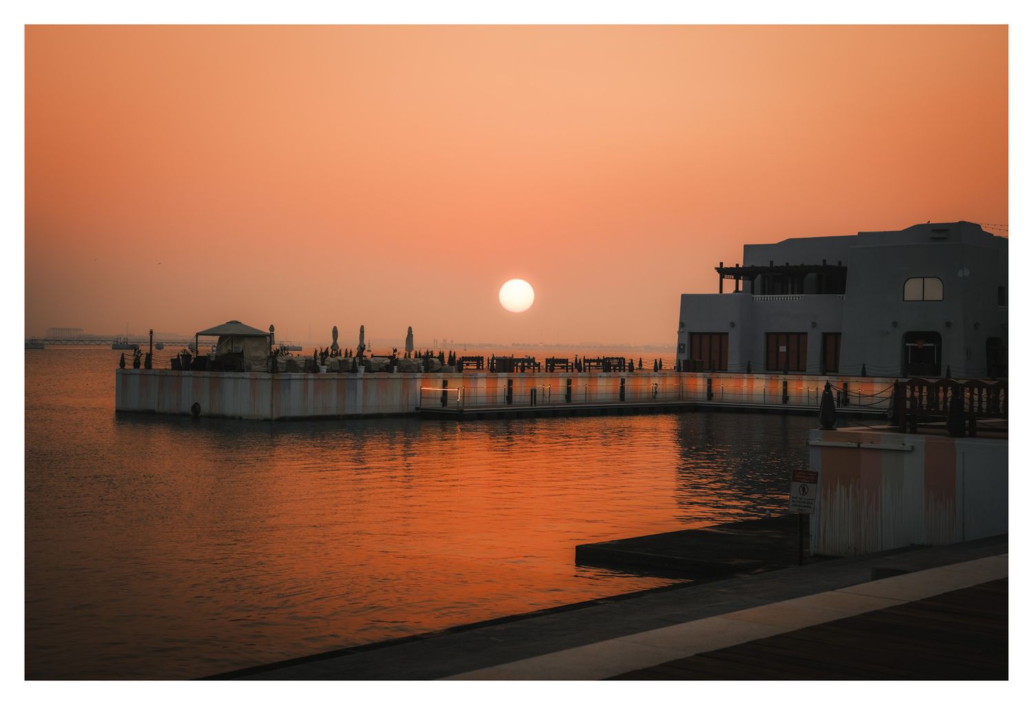 A waterfront pier and buildings silhouetted against a vibrant orange sunset over calm waters.