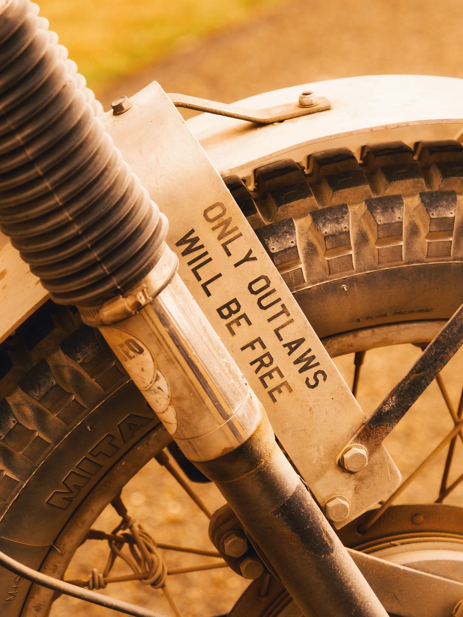 Close-up of a motorcycle fork with a sticker reading 'Only Outlaws Will Be Free' in vintage sepia tone.