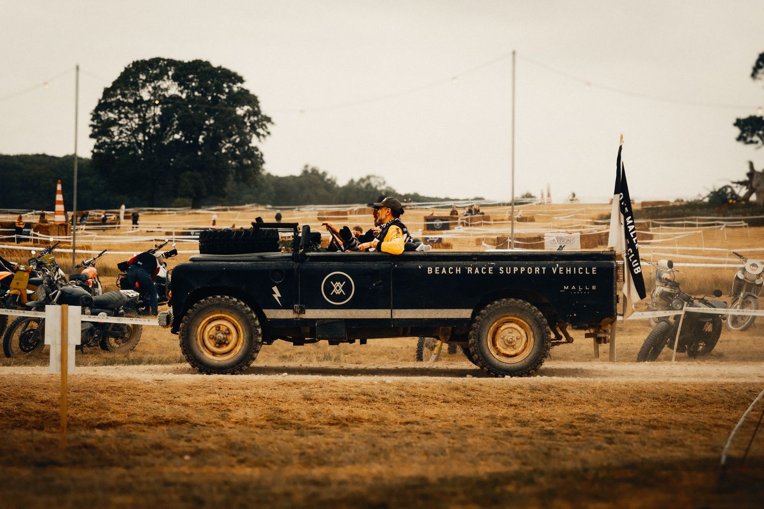 Vintage black Land Rover Beach Race Support Vehicle driving on a dusty field at an outdoor motorsport event.