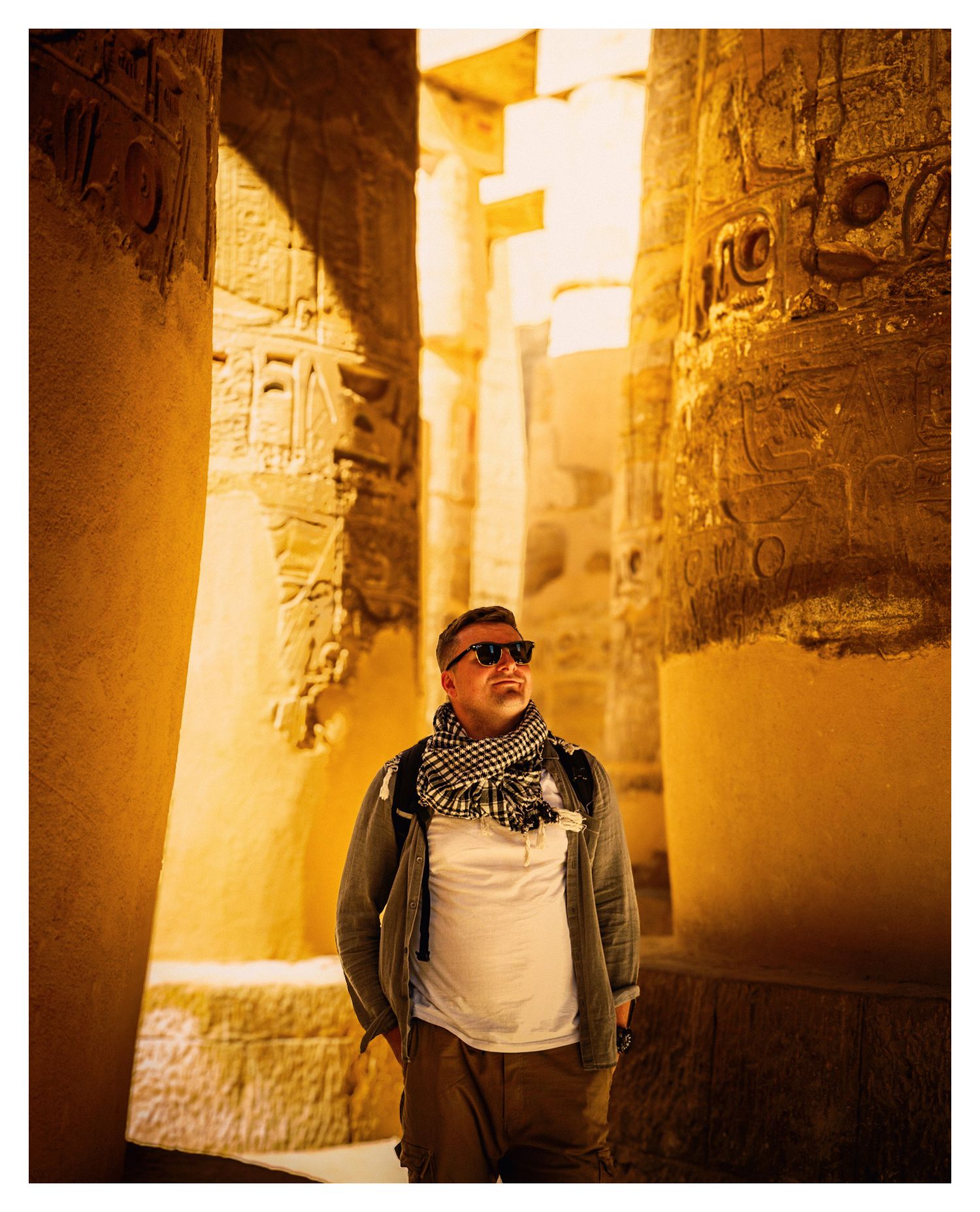 Tourist in sunglasses poses among ancient stone columns at Karnak Temple in warm golden sunlight.