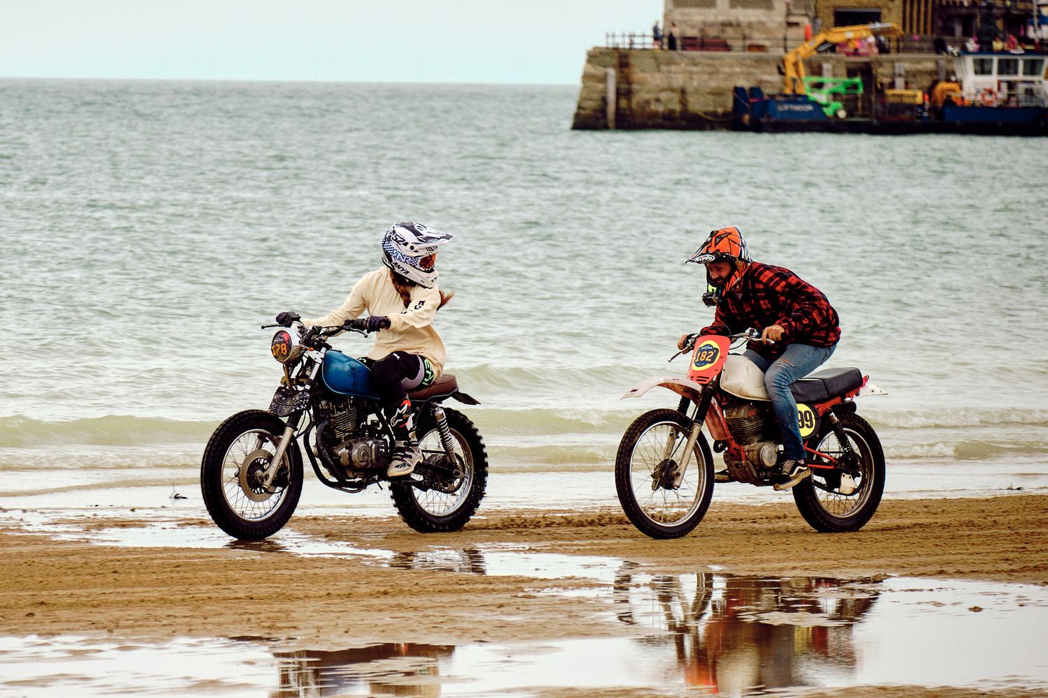 Two motorcycles riding side by side on a sandy beach with water and a coastal structure in the background.