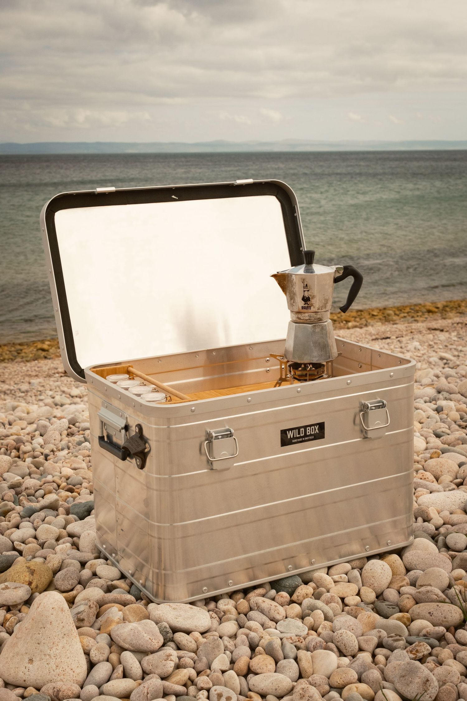 A stainless steel cooler or storage box sits on a rocky beach shoreline with the ocean visible in the background.