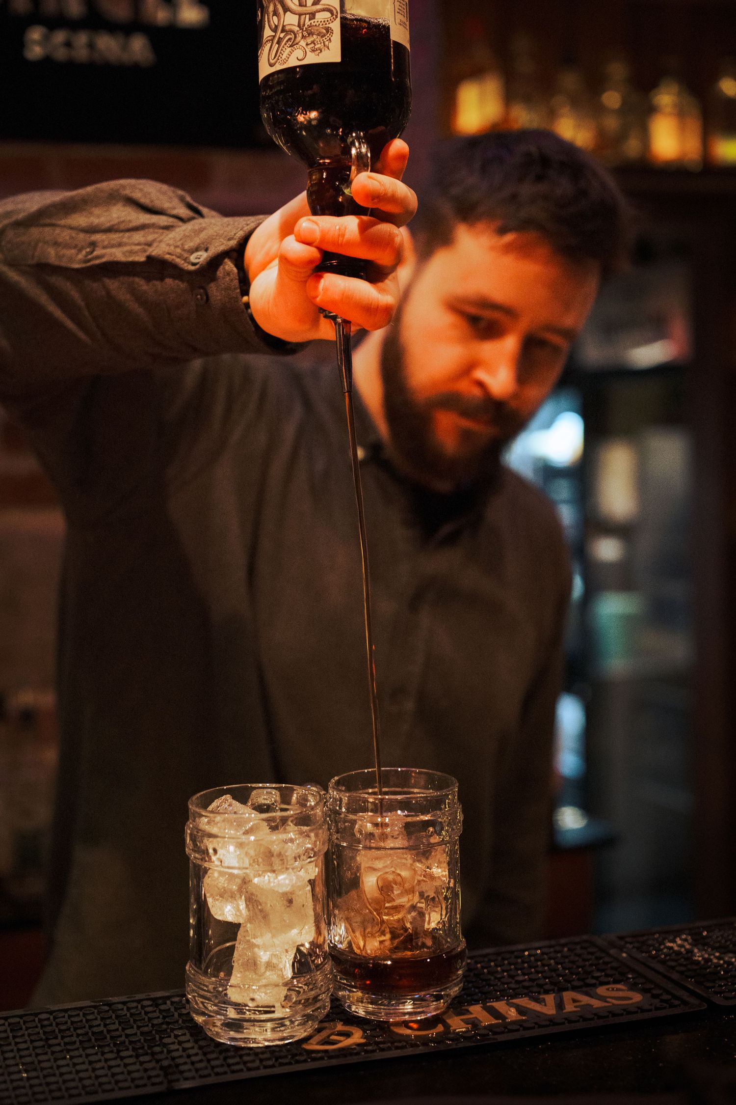 Bartender pours dark liquor into ice-filled rocks glasses at dimly lit bar counter.
