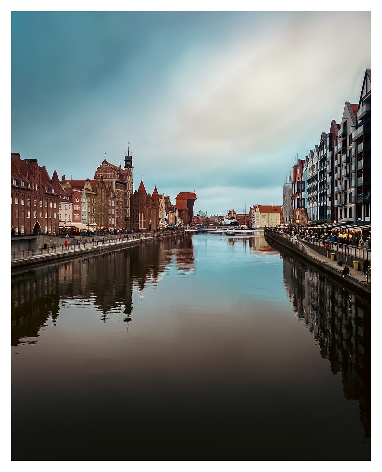 Historic waterfront buildings reflecting in the calm waters of Gdansk's Motlawa River on a cloudy day in Poland.