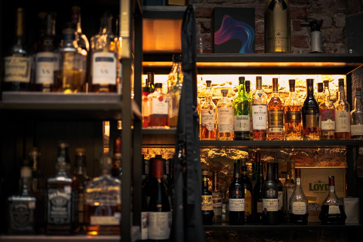Illuminated liquor bottles lined up on glass shelves in a dimly lit bar with warm ambient lighting.