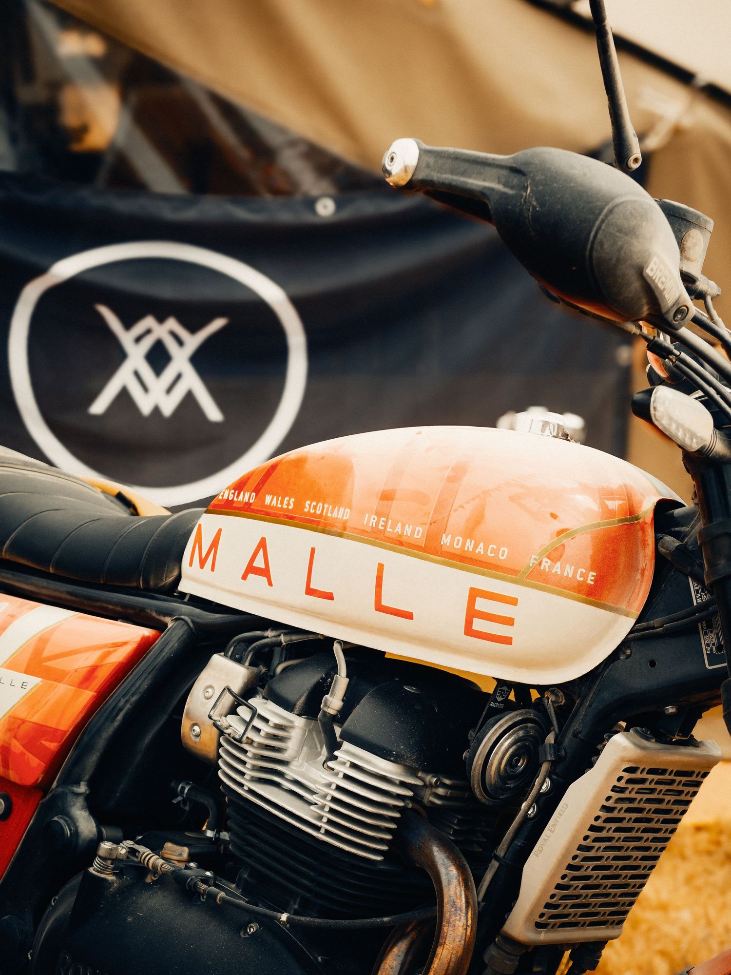 Vintage Malle motorcycle with orange fuel tank displayed outdoors near a branded banner at a rally event.