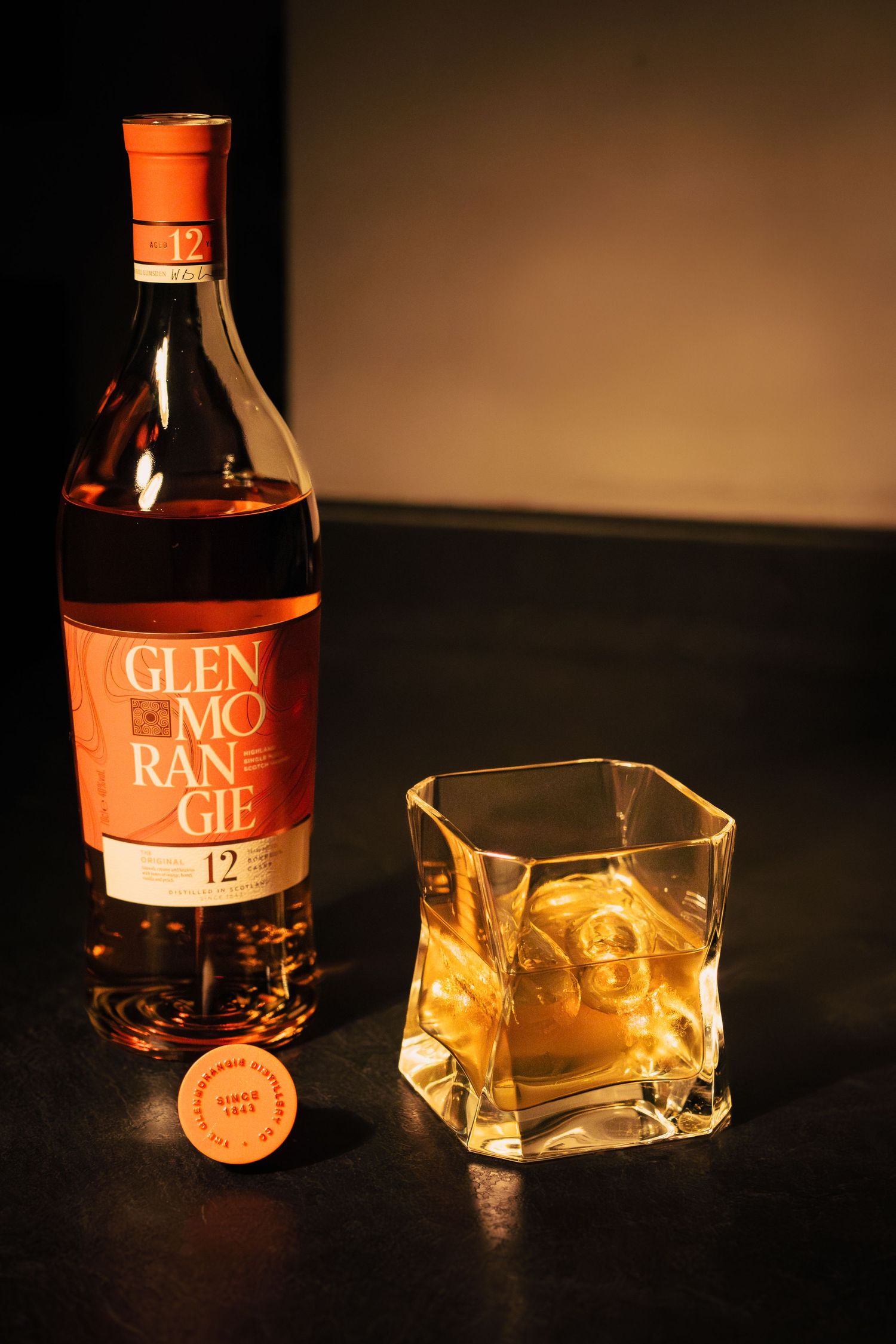 A bottle of Glen Morangie 12 year old whisky alongside a square glass with whisky and ice on a dark surface.
