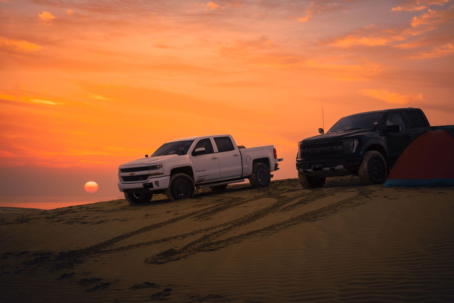 Silhouettes of pickup trucks parked on sand dunes against a vibrant orange sunset sky.