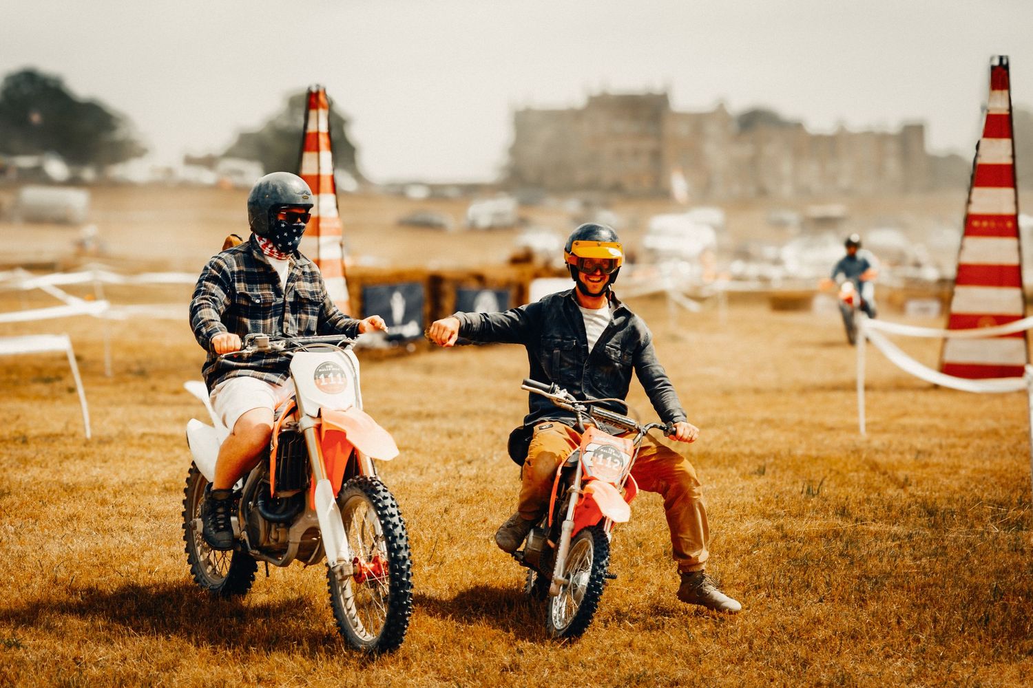Two motocross riders on dirt bikes shaking hands on a grassy field with a castle in the background.