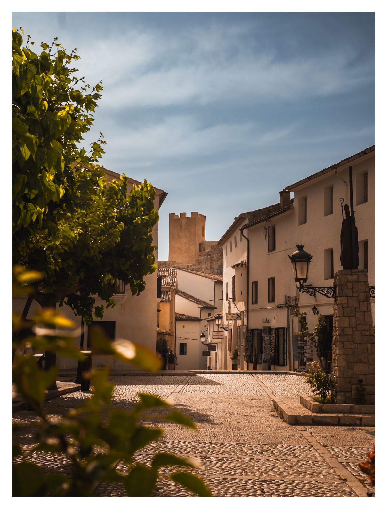 Cobblestone street winding through traditional Spanish village with beige buildings and rustic lampposts under cloudy sky.