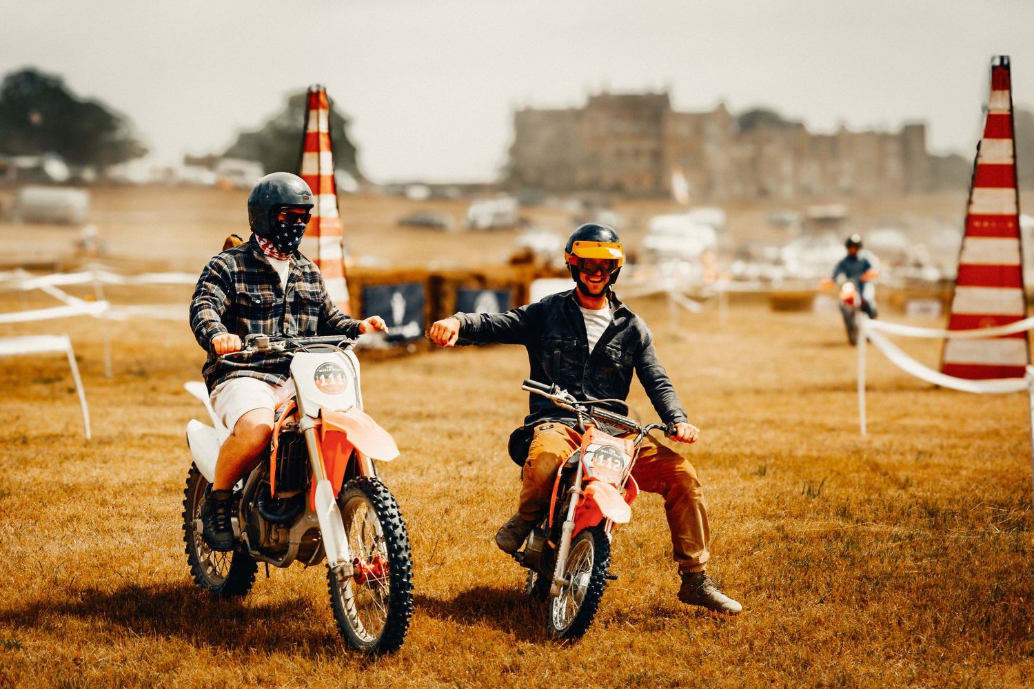 Two motocross riders on dirt bikes shaking hands on a grassy field with a historic building in the background.