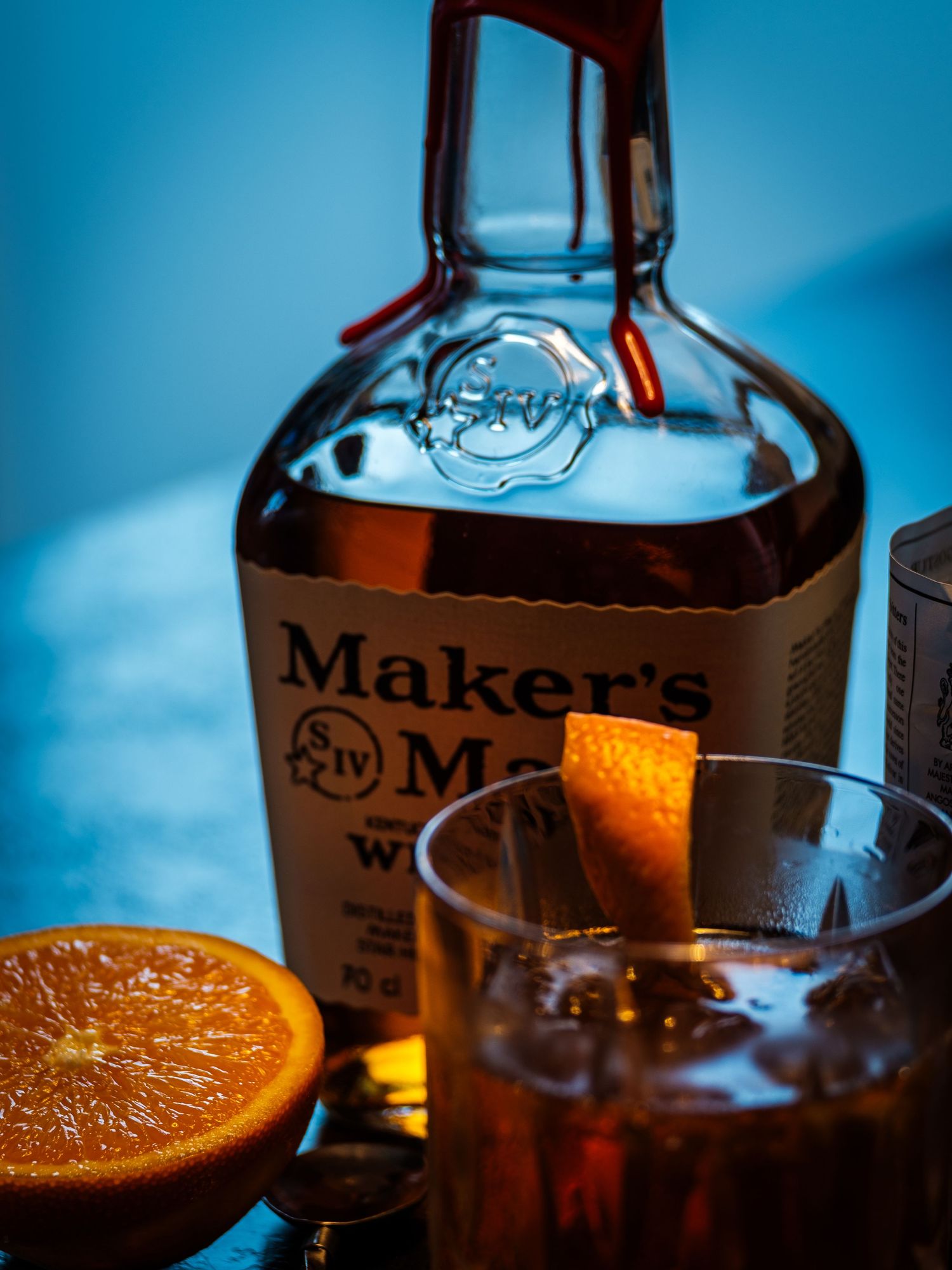 A bottle of Maker's Mark bourbon whiskey with an orange slice and cocktail glass in a moody blue-lit setting.