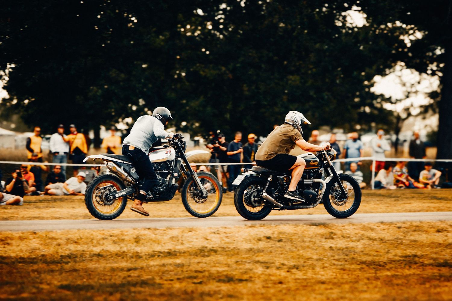 Two motorcyclists race side by side on a dirt track at an outdoor motorsport event with spectators watching.