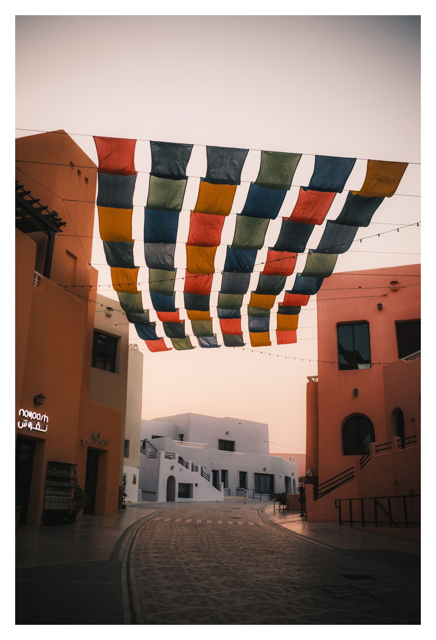 Colorful prayer flags hang between terracotta buildings in a peaceful courtyard at sunset.