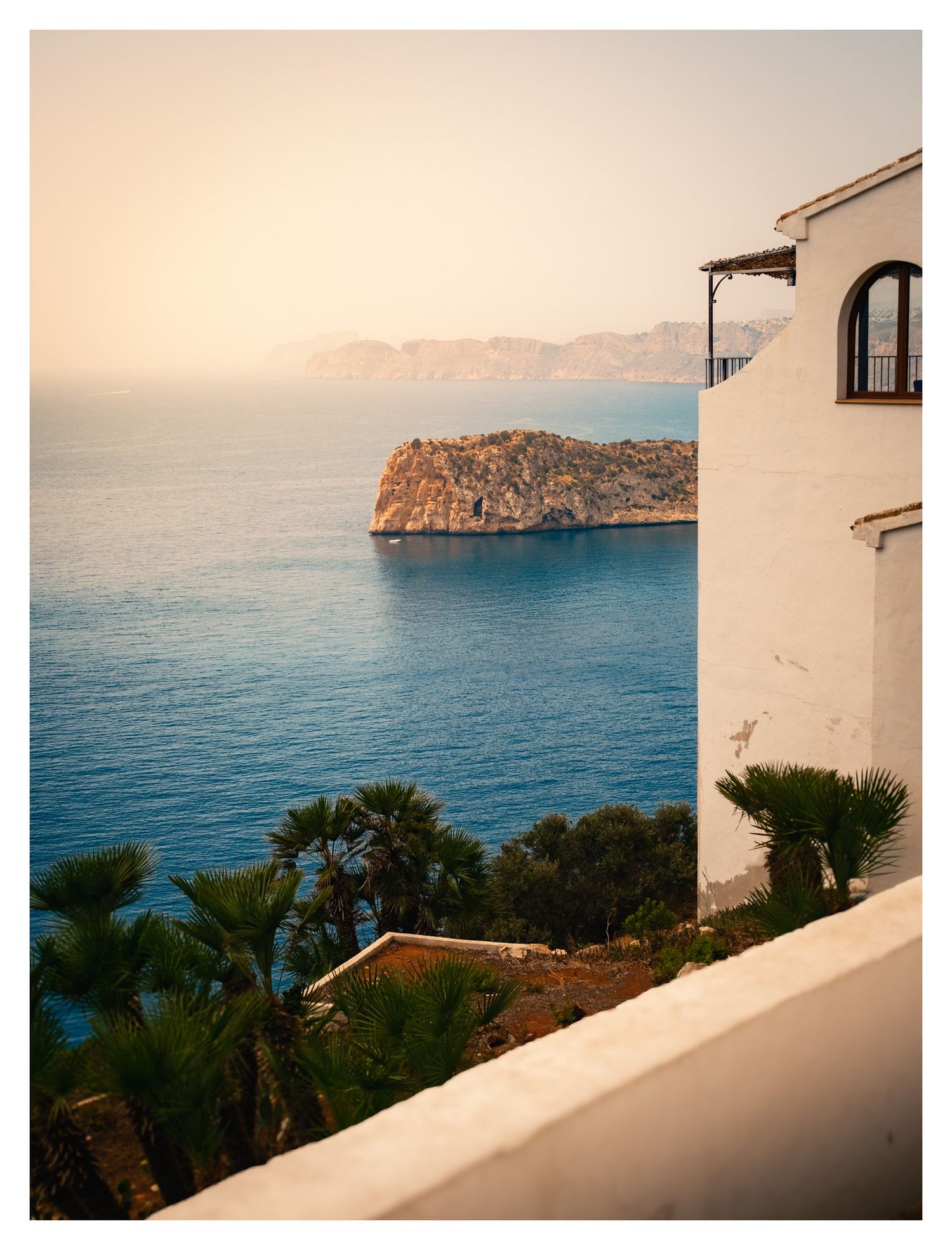 Mediterranean coastal view with a white villa overlooking a rocky island and blue waters during golden hour.