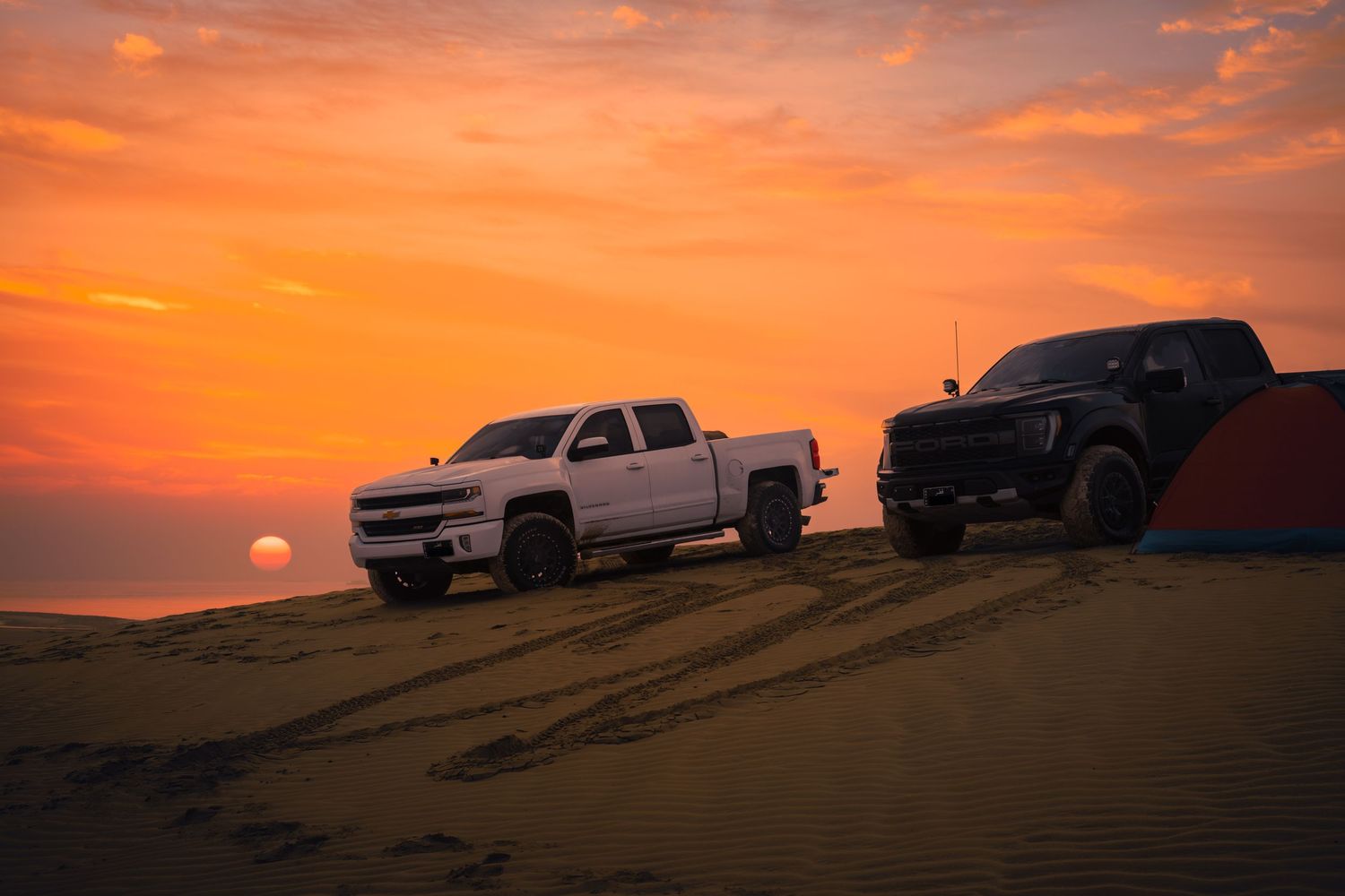 Silhouettes of pickup trucks and SUVs parked on desert sand dunes against a vibrant orange sunrise sky.