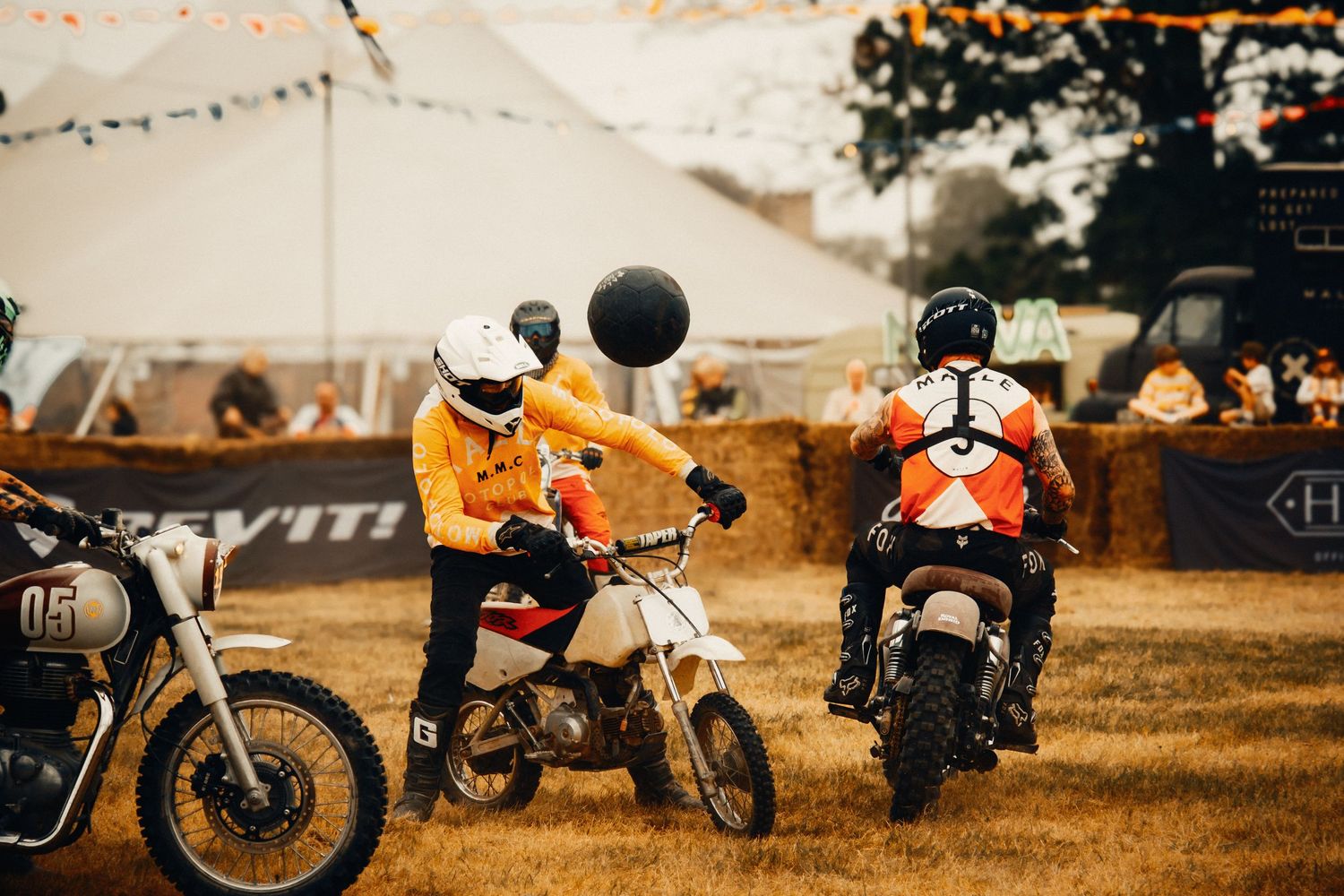 Two riders on dirt bikes play moto polo with a large ball at an outdoor festival event on a dry grass field.