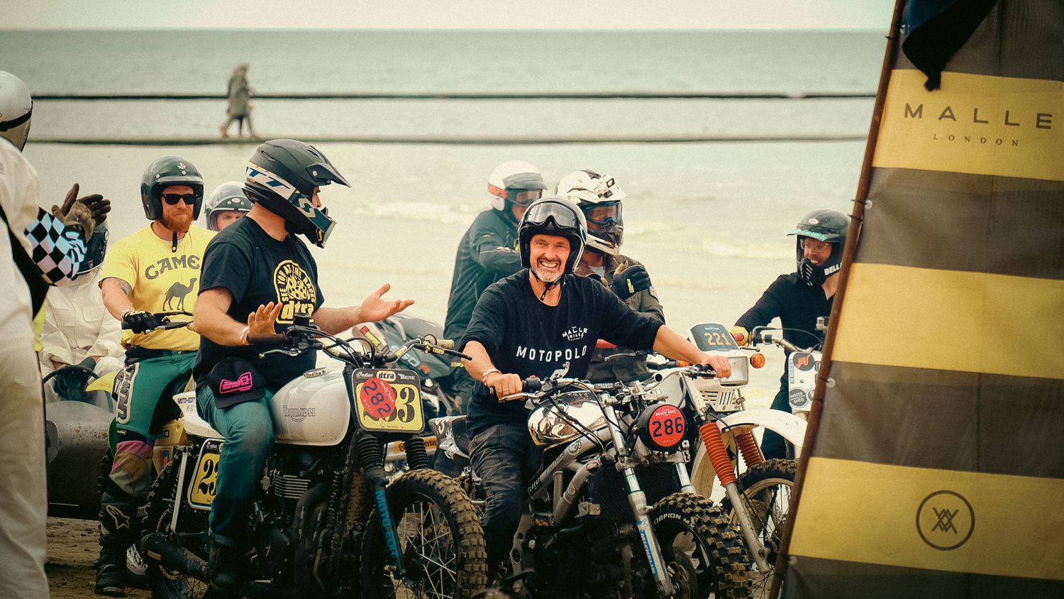 A group of vintage dirt bike riders gathered together on their motorcycles at a muddy outdoor event.