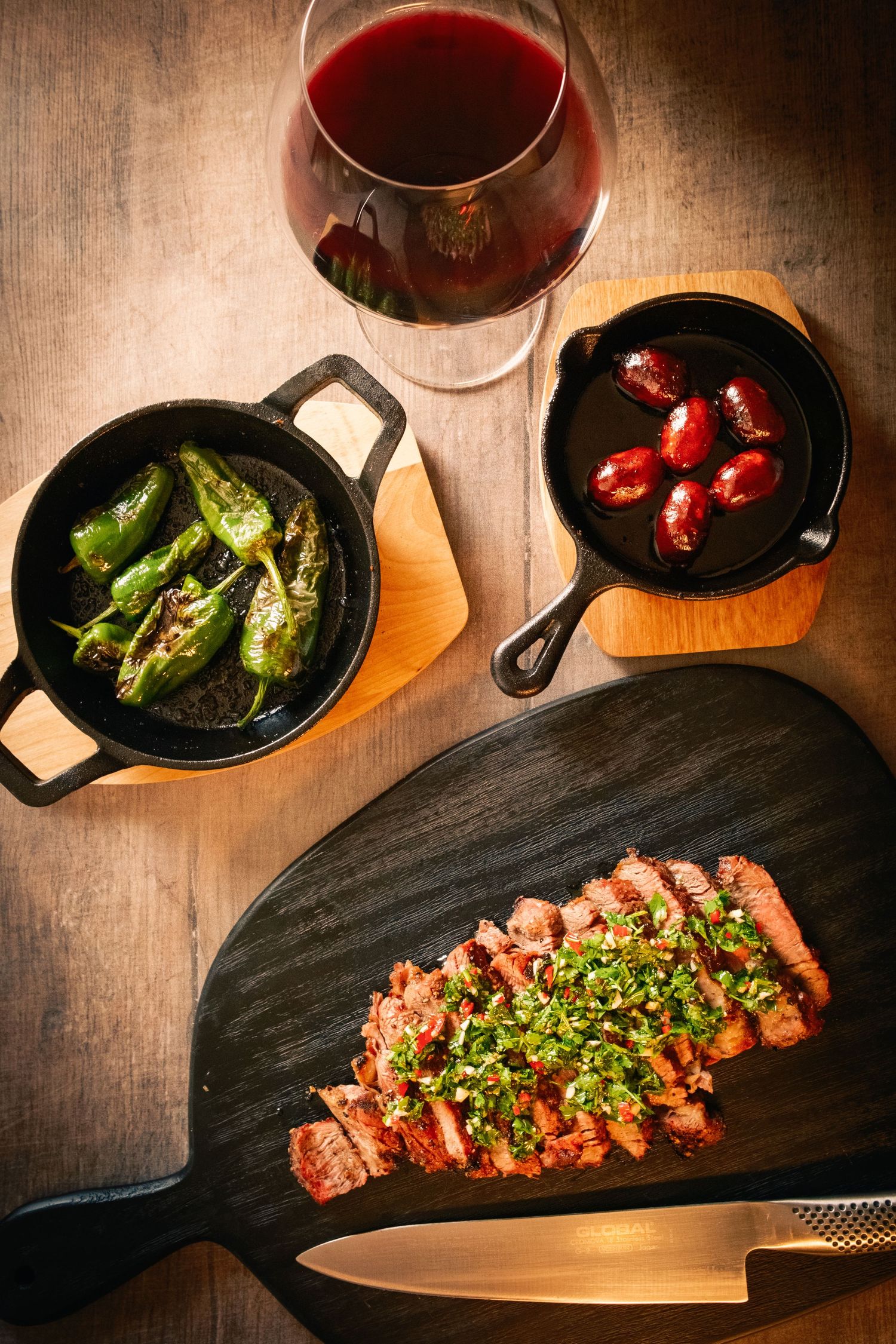 A sequence of wine glasses and cast iron pans with grilled steak and vegetables on a dark wooden surface.
