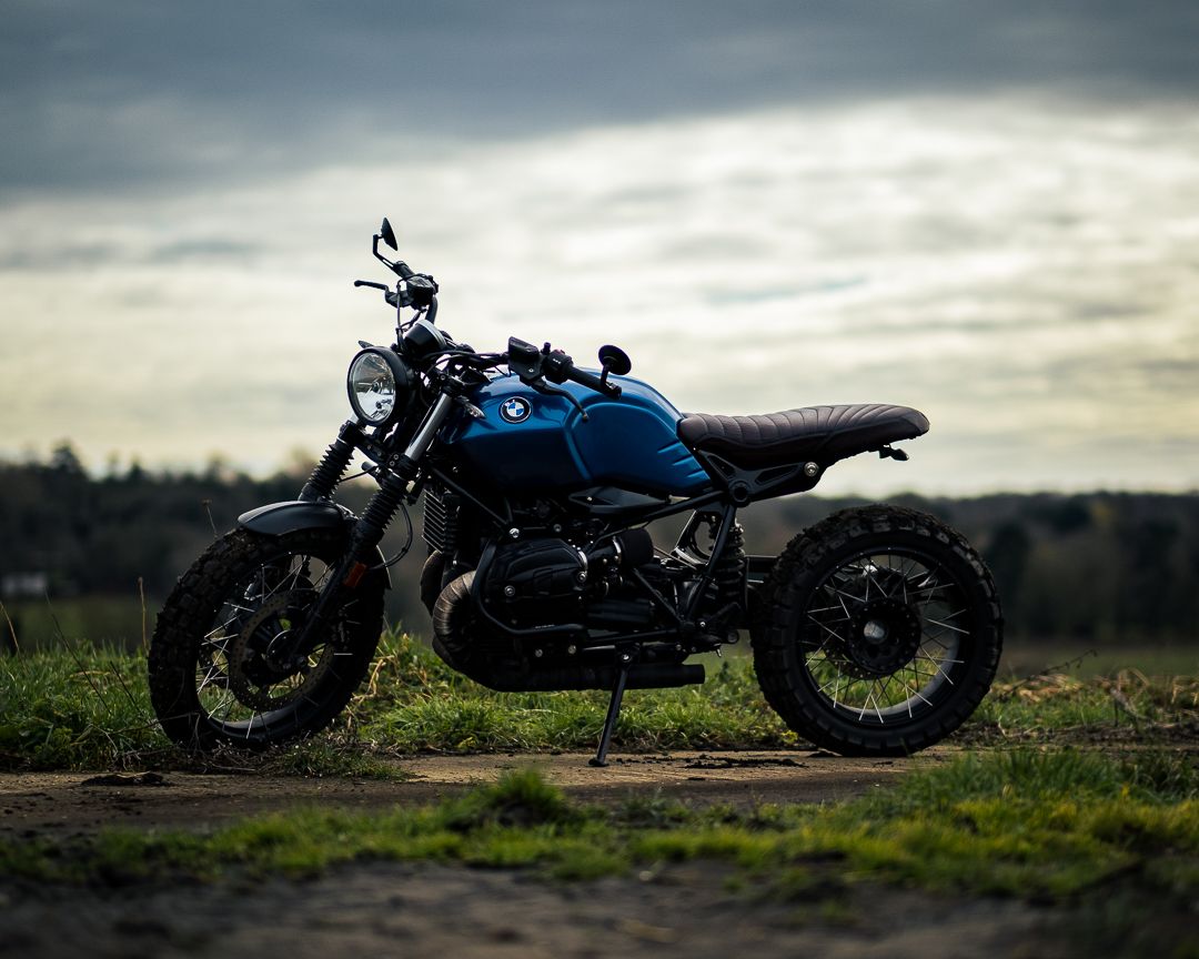 Custom blue motorcycle with sleek cafe racer styling photographed at sunset against dramatic cloudy sky.