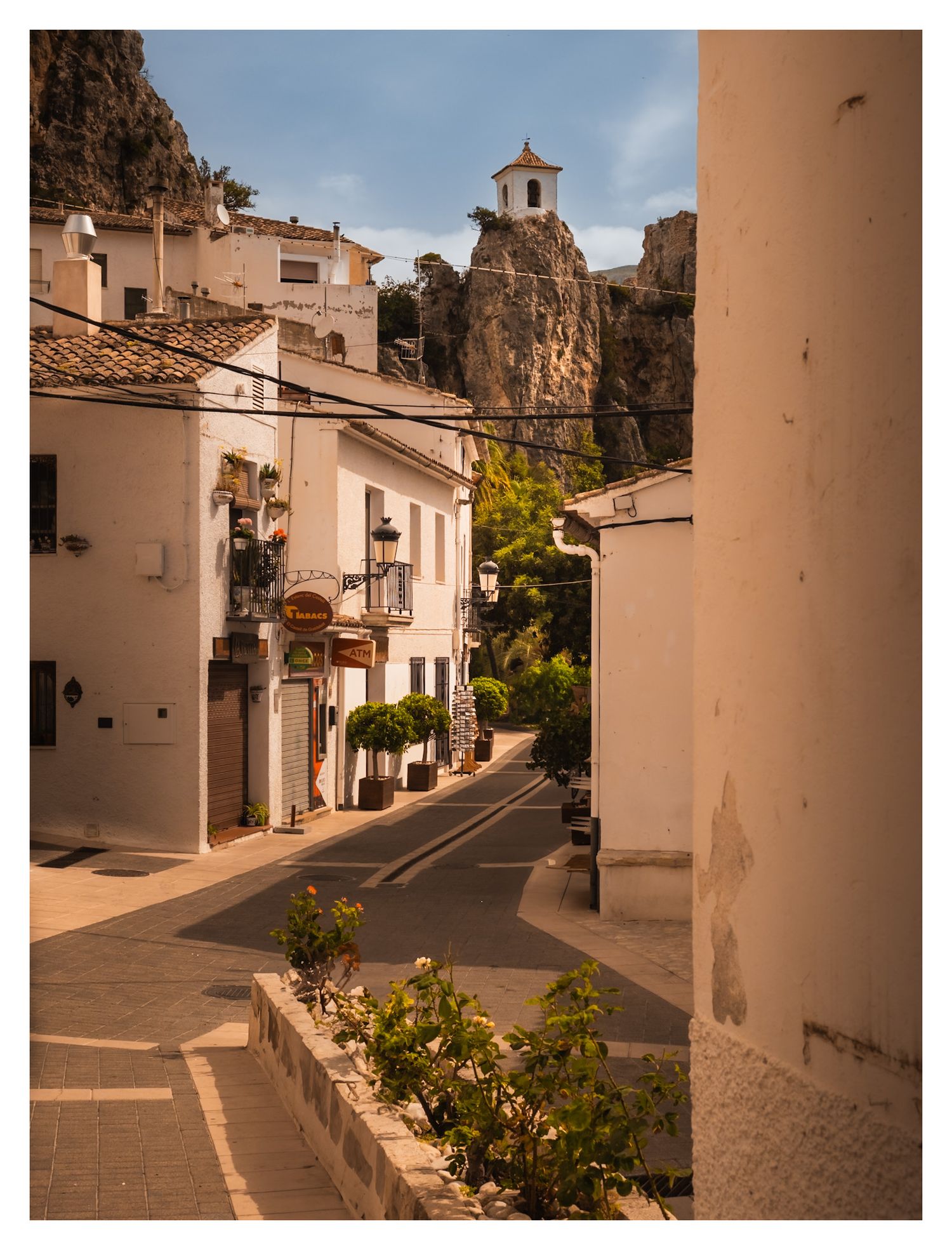 Charming Spanish village street with white buildings and a historic church tower perched atop a rocky hill in the background.