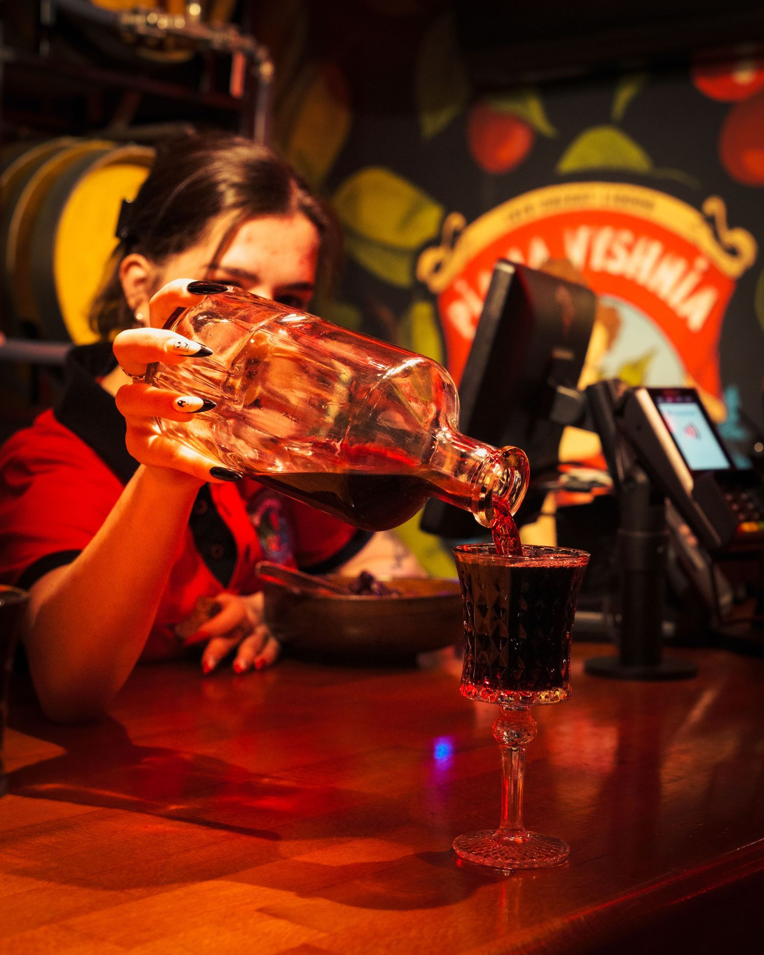 A bartender pours red wine into a glass at a dimly lit bar counter with colorful lights in the background.