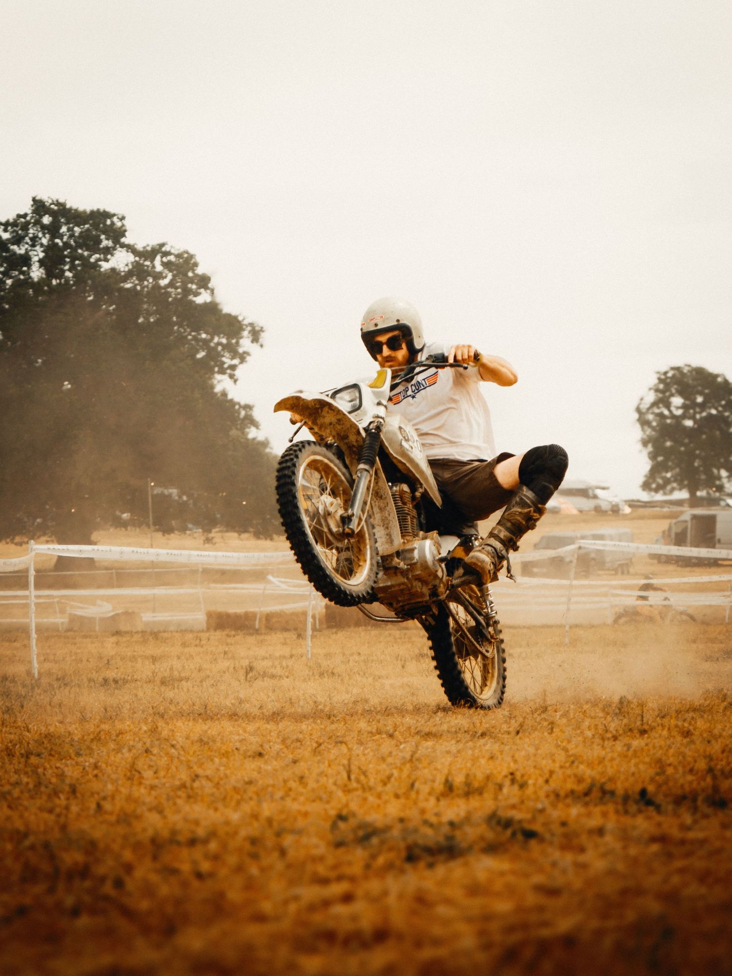 A motocross rider performs a wheelie on a muddy dirt bike at an outdoor track, wearing a white helmet and boots.