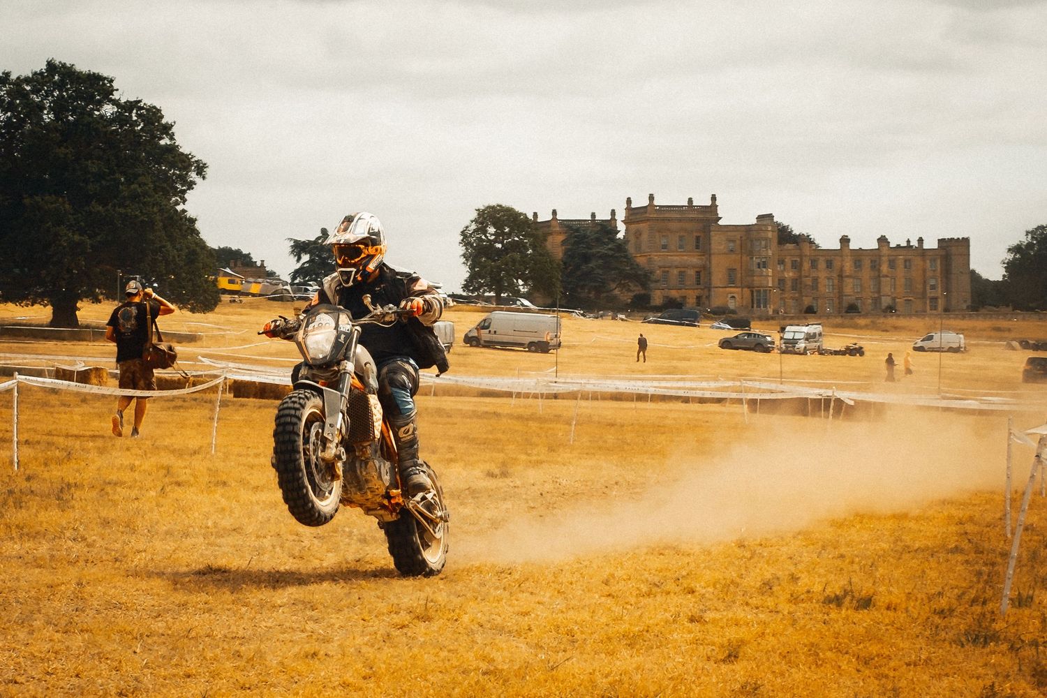 Motorcyclist performing a wheelie on a dirt track at an outdoor event, with a historic mansion visible in the background.
