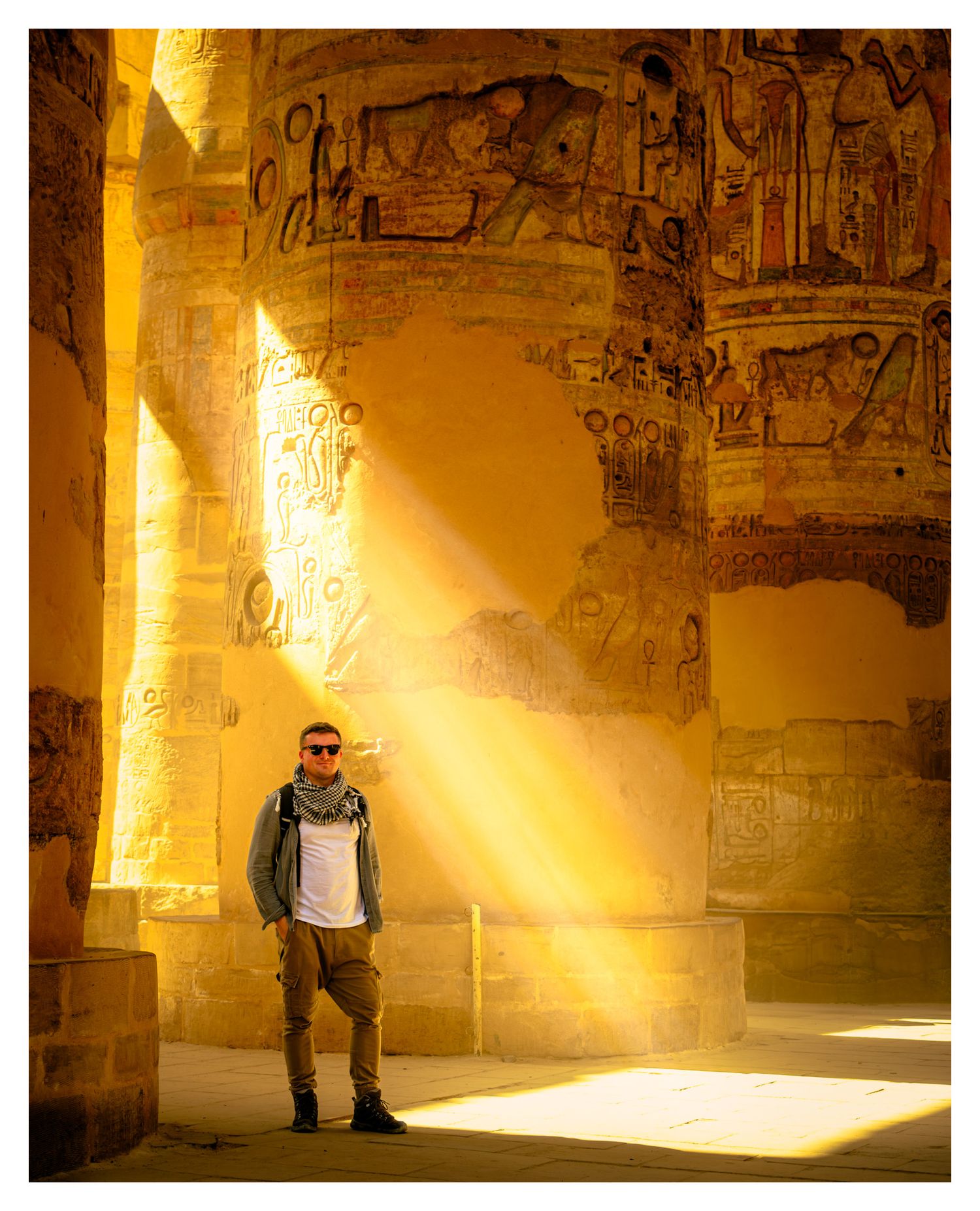 A tourist stands in a beam of sunlight within ancient Egyptian ruins covered in hieroglyphics and carvings.