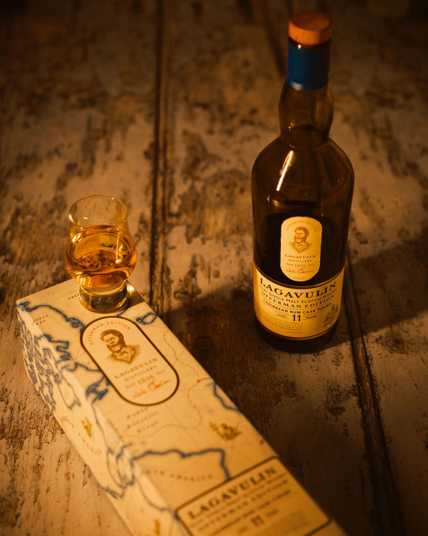 A Lagavulin bottle and glassware arranged on a rustic wooden surface in moody lighting.