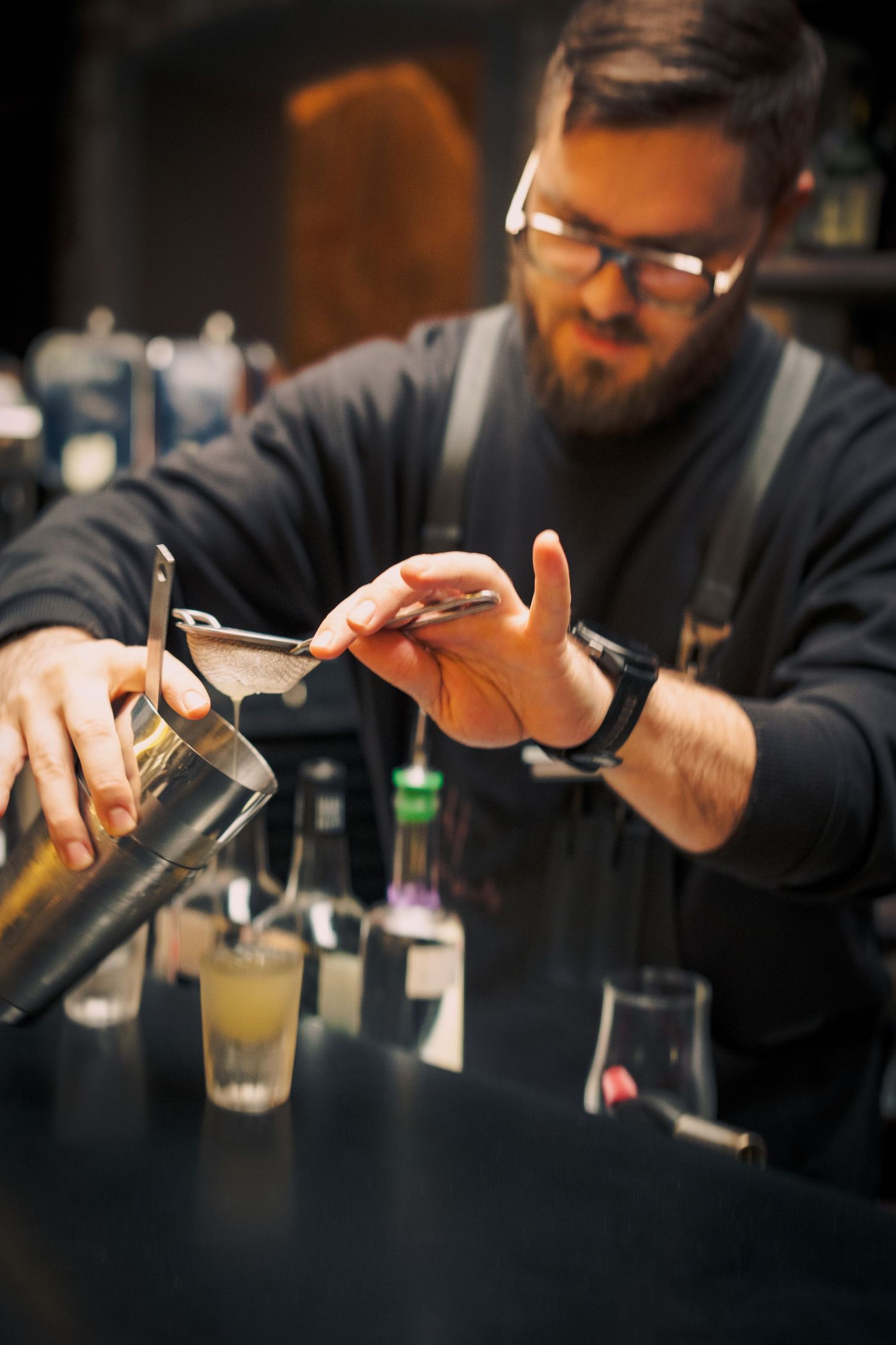 A bartender in glasses and apron pours a drink at a dimly lit bar counter with bottles in the background.