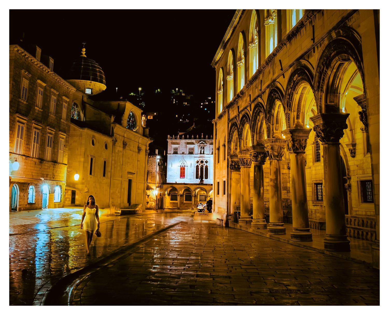 A wet cobblestone street in Dubrovnik's old town at night with illuminated historic arches and buildings.