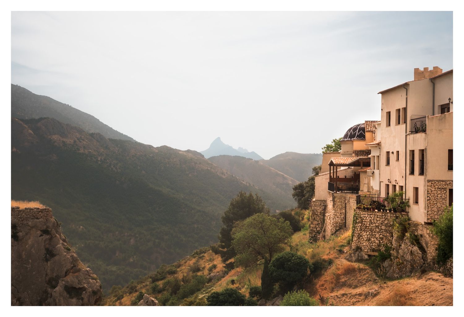 Traditional stone buildings perch on rocky cliffs overlooking a mountainous valley in Guadalest at sunset.